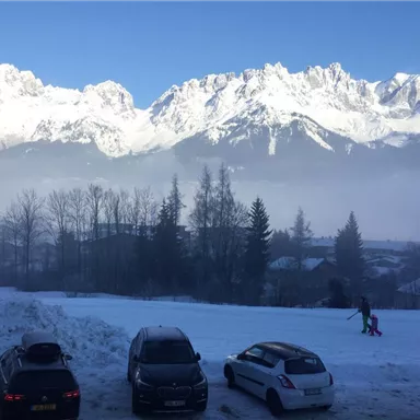 Eine schneebedeckte Landschaft mit majestätischen Bergen im Hintergrund. Im Vordergrund stehen einige Autos und eine Person, die im Schnee spaziert.
