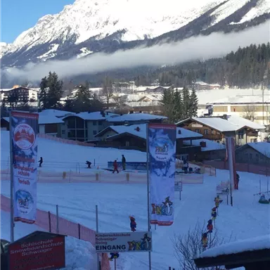Eine malerische Winterszene mit schneebedeckten Bergen im Hintergrund. Auf der Piste sind Menschen beim Skifahren und Snowboarden zu sehen.