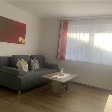 A cozy living room with a gray sofa and a table. The windows are bright and decorated with sheer curtains.