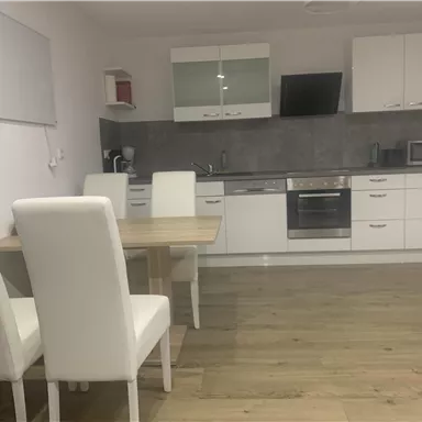A modern kitchen with white furniture and wooden flooring. Four white chairs stand at the table, and the kitchen setup is simple and functional.