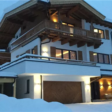 Ein modernes Chalet im Schnee, umgeben von einer winterlichen Landschaft. Die Fassade ist hell und strukturiert, mit einem großen Balkon und warmem Licht.
