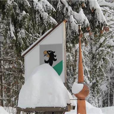 Ein Schild mit einem Wappen und einem schwarzen Adler, umgeben von Schnee. Im Hintergrund sind schneebedeckte Bäume zu sehen.