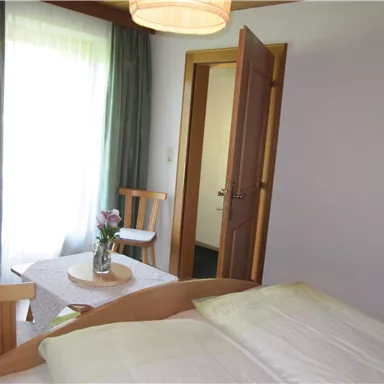 Ein helles Zimmer mit einem Bett, einem Tisch und Stühlen. Das Fenster lässt viel Licht herein und schafft eine einladende Atmosphäre.