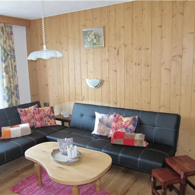 Ein gemütliches Wohnzimmer mit Holzverkleidung und zwei schwarzen Sofas. In der Mitte steht ein runder Tisch mit dekorativen Elementen und bunten Kissen auf den Sofas.