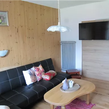 Ein gemütliches Wohnzimmer mit einem schwarzen Sofa und bunten Kissen. An der Wand hängt ein Fernseher und neben dem Sofa steht ein runder Tisch.