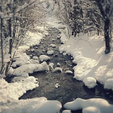 A narrow stream flows through a snow-covered landscape. The banks are covered with fresh snow and round stones.