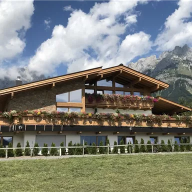 Ein modernes, gut gestaltetes Haus mit einem Balkon voller Blumen. Im Hintergrund sind majestätische Berge und ein blauer Himmel mit einigen Wolken zu sehen.