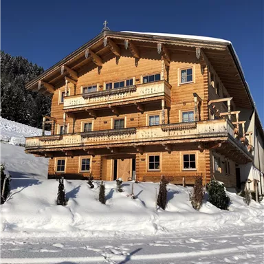 A large wooden house in the snow with several windows and balconies. The surroundings are wintry with snow-covered trees.