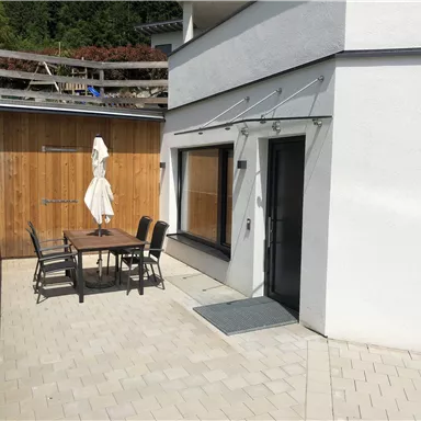 Ein modernes Haus mit einem gepflasterten Außenbereich. Ein Esstisch mit Stühlen und ein Sonnenschirm stehen bereit.