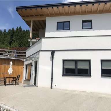 Ein modernes, zweigeschossiges Haus mit weißer Fassade und Holzdetails. Vor dem Haus befindet sich eine Terrasse mit einem Tisch und Stühlen.