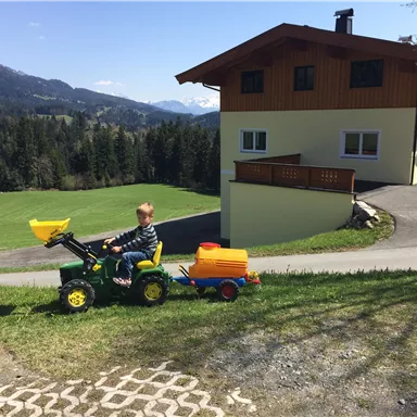 Ein kleines Kind fährt mit einem Spielzeugtraktor auf einem Hügel. Im Hintergrund ist ein schönes Haus und eine grüne Wiese.