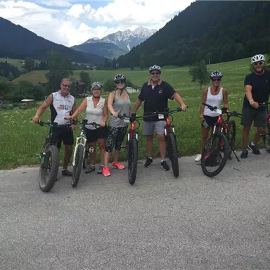 A group of six people is standing with bicycles on a path in a picturesque landscape. In the background, green hills and mountains can be seen.