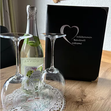 A bottle of drink with two glasses on a table, dressed with a small lace coaster. In the background, there is a welcome sign in a black folder.