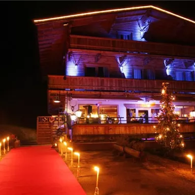 Ein gemütliches Bergchalet bei Nacht, beleuchtet mit bunten Lichtern. Ein roter Teppich führt zur Eingangstür, umgeben von Fackeln und festlicher Dekoration.