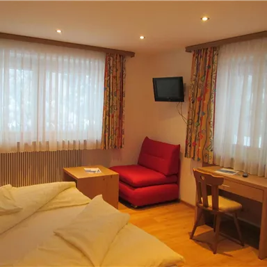 A cozy room with a double bed and a red armchair. Large windows with curtains let in plenty of light.