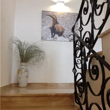 Ein einladender Treppenaufgang mit Holzstufen und einem dekorativen Geländer. An der Wand hängt ein Bild eines Bergtieres, und in der Ecke steht eine Vase mit grünem Dekor.