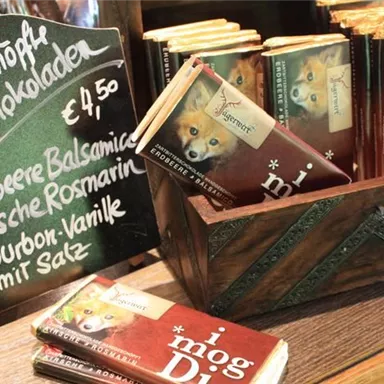 Ein rustikaler Holzstand mit verschiedenen Schokoladentafeln. Neben den Schokoladen steht eine Tafel mit dem Preis und den angebotenen Geschmacksrichtungen.