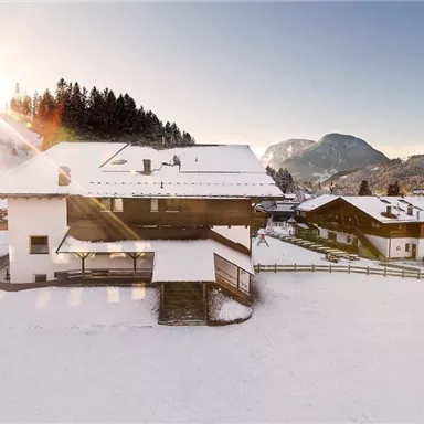 Ein wunderschönes Winterlandschaft mit schneebedeckten Häusern und einem klaren blauen Himmel. Die Sonne scheint über die Berge und verleiht der Szene eine malerische Atmosphäre.