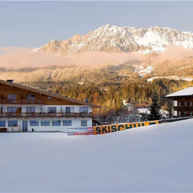 Eine verschneite Berglandschaft mit zwei Chalet-Häusern. Im Hintergrund erstrecken sich schneebedeckte Berge und ein klarer Himmel.