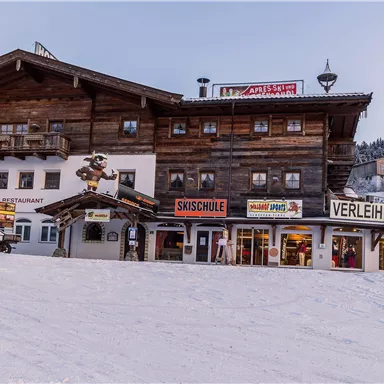 Ein traditionelles Holzgebäude im Schnee mit Beschilderungen für Skischule und Verleihservice. Die Umgebung ist winterlich und einladend für Wintersportler.