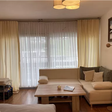 A cozy living room with a bright sofa and a wooden table. Large windows with white curtains provide plenty of daylight.