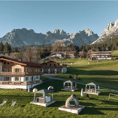 Ein malerisches Alpenresort mit traditionellen Holzgebäuden und einer grünen Wiese. Im Hintergrund erheben sich beeindruckende Berge unter klarem Himmel.