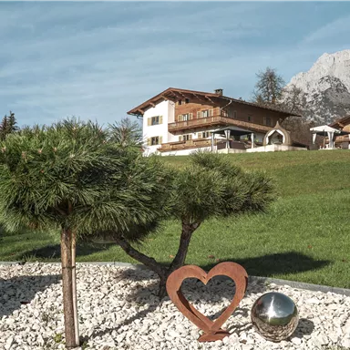 Ein schönes Haus in den Bergen umgeben von grünem Rasen und einem gepflegten Garten. Im Vordergrund steht eine herzförmige Skulptur neben einer glatten Metallkugel.