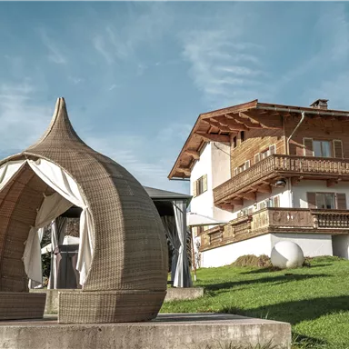 Ein moderner Pavillon aus geflochtenem Material steht auf einer Wiese. Im Hintergrund ist ein traditionelles Holzhaus mit Balkonen zu sehen.