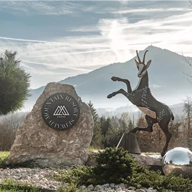 Ein stilisiertes Antilopen-Symbol steht vor einer markanten Berglandschaft. Die Umgebung ist grün mit Sträuchern und einem klaren Himmel.
