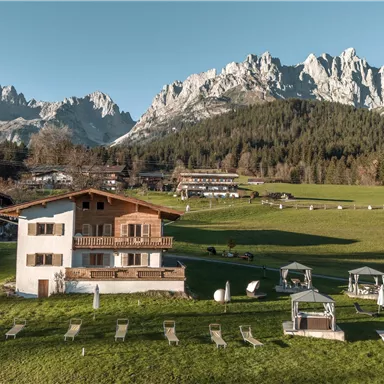 Eine malerische Berglandschaft mit einem modernen Chalet und traditionellen Hütten. Der grüne Rasen und die beeindruckenden Berge im Hintergrund schaffen eine idyllische Atmosphäre.