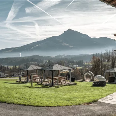 Eine malerische Landschaft mit Bergen im Hintergrund und Nebel über dem Tal. Im Vordergrund stehen gemütliche Pavillons und eine gepflegte Wiese.