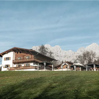 Ein traditionelles Haus inmitten einer grünen Wiese, mit beeindruckenden Bergen im Hintergrund. Der Himmel ist klar und die Landschaft wirkt friedlich.