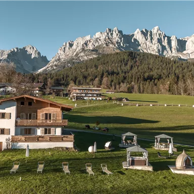 Eine malerische Landschaft mit Bergkulisse und einem gemütlichen Haus. Im Vordergrund sind Liegestühle und Gartenmöbel zu sehen.