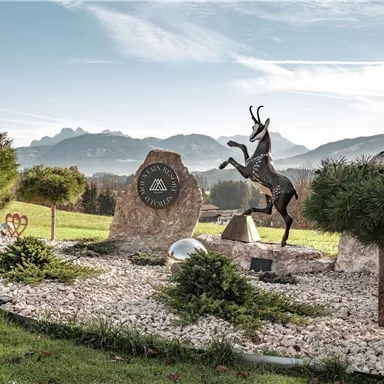 Ein schöner Garten mit Steindenkmälern und einer Statue eines reitenden Steinbocks. Im Hintergrund sind die Berge und ein klarer Himmel sichtbar.