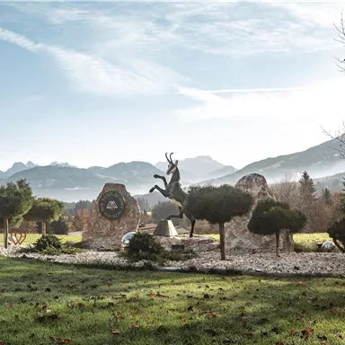 Eine ruhige Landschaft mit sanften Hügeln und klarer Bergsicht. Im Vordergrund steht ein dekoratives Element mit einem Hirsch und gepflegten Bäumen.