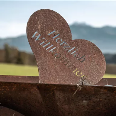 Ein herzförmiger Willkommensschild aus Metall steht auf einem Gartenbeet. Im Hintergrund sind sanfte Hügel und Berge zu sehen.