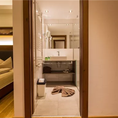 Ein modernes Badezimmer mit einer Glasdusche und hellen Fliesen. Im Hintergrund ist ein gemütliches Schlafzimmer mit einem großen Bett zu sehen.