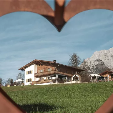 Ein gemütliches Chalet umgeben von grünem Gras und Bergen. Der Blick wird durch eine herzförmige Öffnung gerahmt.