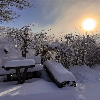 Eine verschneite Winterlandschaft mit einem Tisch und einer Bank, die von Schnee bedeckt sind. Im Hintergrund ist die Sonne, die über den Horizont scheint.