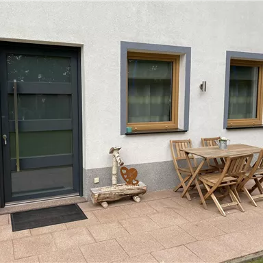 Ein gemütlicher Innenhof mit einem Holzstammtisch und Stühlen. Neben dem Tisch steht ein Grill und die Fenster sind mit grünen Vorhängen ausgestattet.