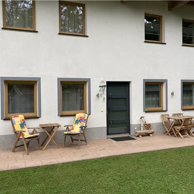 Ein gemütliches Haus mit hellem Anstrich und großen Fenstern. Draußen stehen zwei bunte Liegestühle und ein kleiner Tisch auf einer Terrasse.