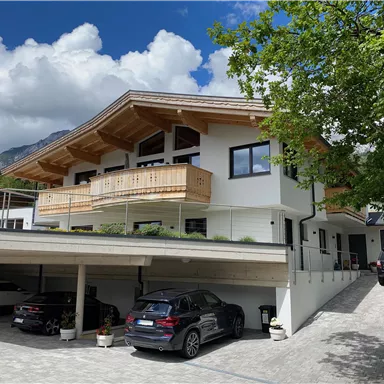 A modern house with a wooden balustrade and a covered garage. In the background, blooming trees and a blue sky can be seen.