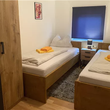 A cozy room with two single beds and wooden furniture. On the beds are yellow towels, and the window is covered with a dark roller blind.