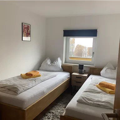 A simple bedroom with two single beds and white bedspreads. On the beds are orange towels, and a window lets in daylight.
