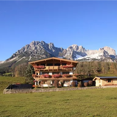 Ein schönes Haus mit Holzveranda in einer malerischen Berglandschaft. Im Hintergrund sind beeindruckende Berge und ein blauer Himmel zu sehen.