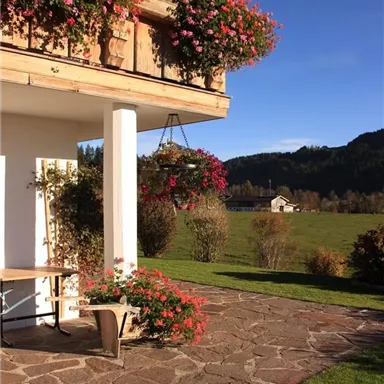 Ein schöner Eingangsbereich mit blühenden Pflanzen und einer steinernen Terrasse. Im Hintergrund sind sanfte Hügel und ein blaues Himmel sichtbar.