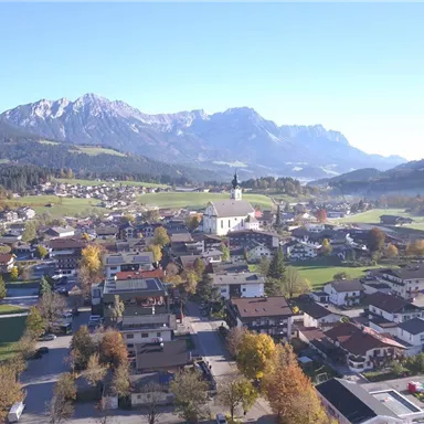 Eine malerische Dorfansicht mit traditionellen Häusern und einer Kirche im Vordergrund. Im Hintergrund erheben sich majestätische Berge unter einem klaren Himmel.