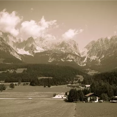An impressive mountain landscape with high peaks and a clear sky. In the foreground, a green meadow stretches out along with small buildings.