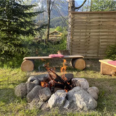 A cozy campfire, surrounded by stones and wooden benches. In the background, trees and a wooden wall are visible.