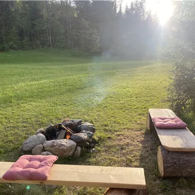 A cozy campfire area with a wooden bench and cushions. Surrounded by a green meadow and trees in the background.